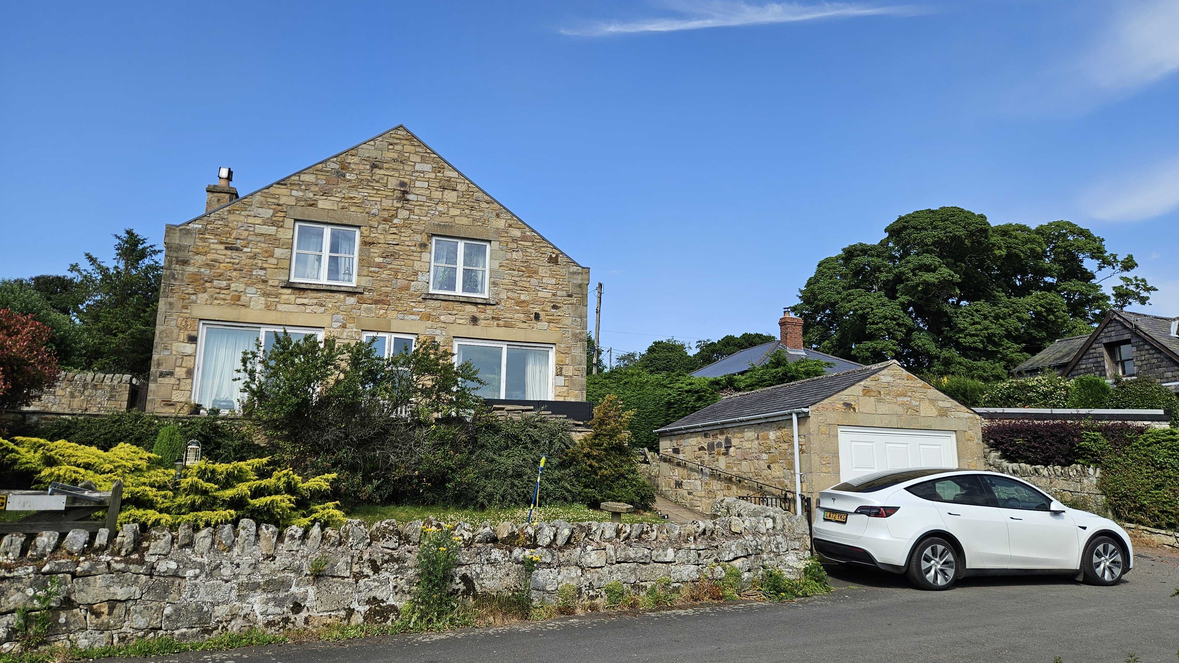 A stone-built house with a garden, a low stone wall, and a car parked by the garage