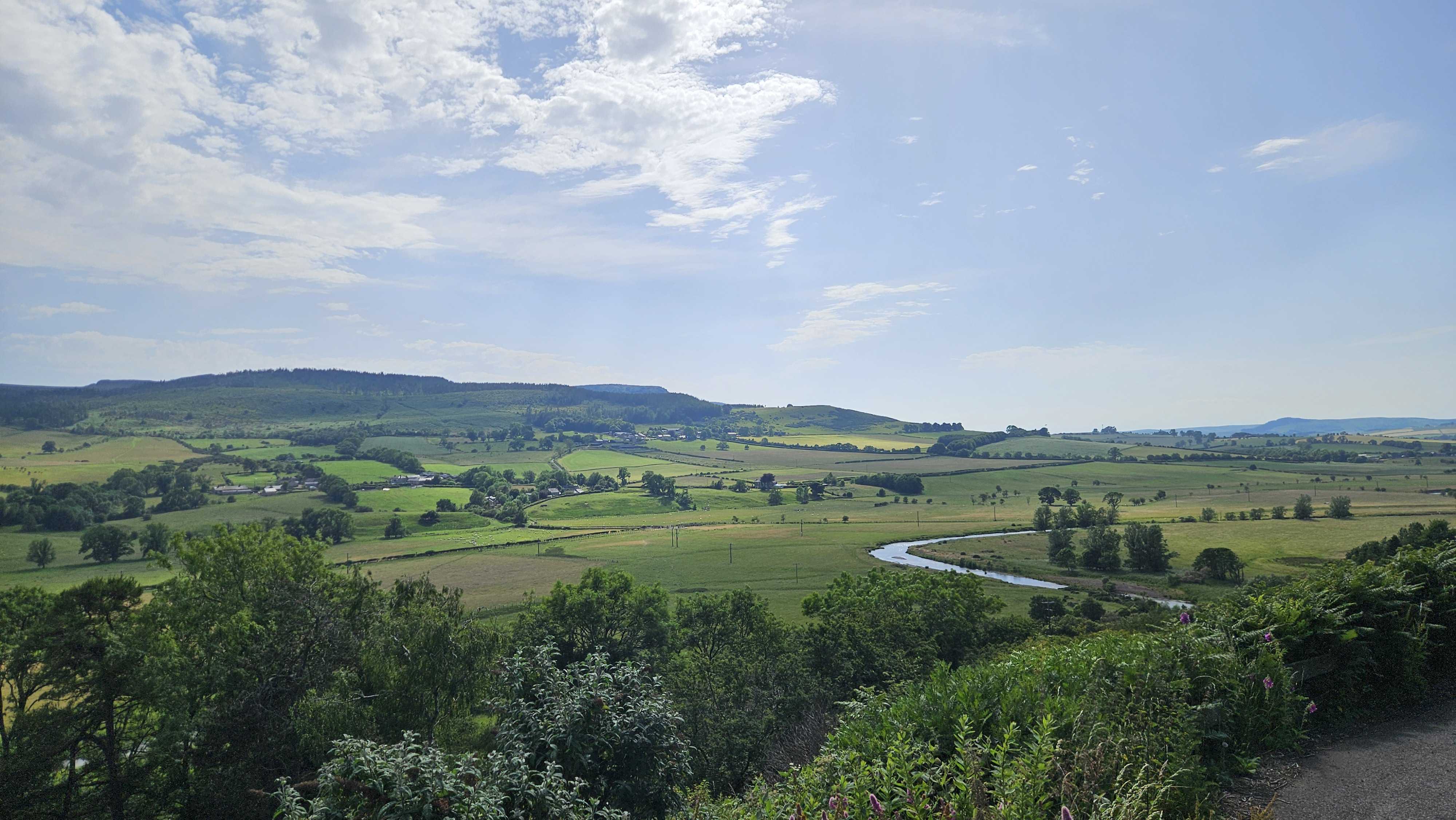 A wide view over green rolling countryside, with fields, hills, and a river winding through the valley